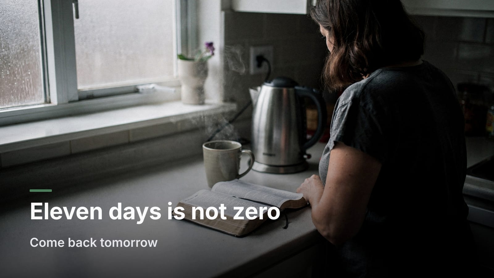 Woman seen from behind reading an open Bible at a kitchen counter beside a mug and kettle in morning light